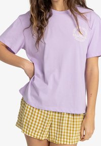 En kvinna som bär en ljuslila Billabong-t-shirt och gula rutiga shorts, med en hand vilande på höfterna mot en vit bakgrund.