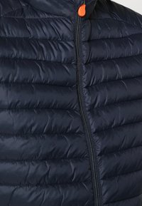Marineblå puffervest med quiltet tekstur, frontlynlås i sølvfarve og orange lynlåsdrage. Har et let og isoleret design.