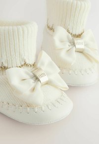 Valged beebikummikud ribakudumistega ääres, kangast paeltega ja hõbedase logosildiga. Omab õmblusdetalje ja pehmeid tallasid.