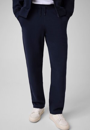 Persona con pantalones azul marino con cordón, zapatillas blancas y una chaqueta azul marino parcialmente visible con las manos en los bolsillos.