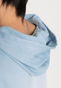 Sudadera de un azul claro con una textura suave, que cuenta con capucha ajustable y costuras visibles. El escote muestra una vista incompleta de la prenda.