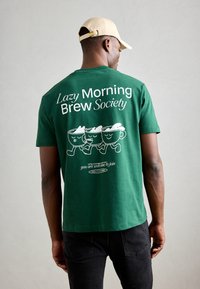 Grønn bomull t-skjorte med tre tegneserie kaffekopper, tekst "Lazy Morning Brew Society," og et tillegg av en kaffe-relatert slogan nedenfor.