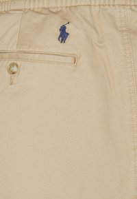 Beige shorts av mjukt material; har en framficka, knäppning med knapp och en blå broderad logotyp av en polo-spelare på sidan.