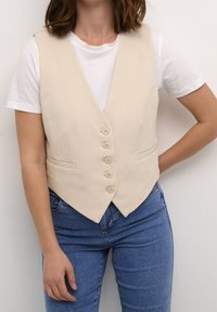 Gilet beige senza maniche con scollo a V, sei bottoni sul davanti e due tasche laterali, indossato sopra una maglietta bianca e abbinato a jeans.