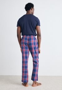 T-shirt marine et pantalon de pyjama à carreaux rouges, bleus et blancs avec une taille élastique. Modèle montré de dos, pieds nus sur un sol moquetté.