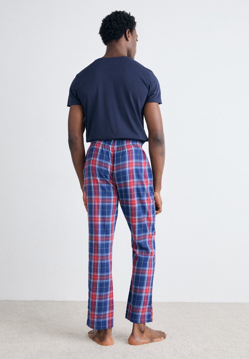 Marineblaues T-Shirt und rot, blau und weiß karierte Schlafanzughose mit elastischem Bund. Das Modell wird von hinten gezeigt und steht barfuß auf einem mit Teppich ausgelegten Boden.