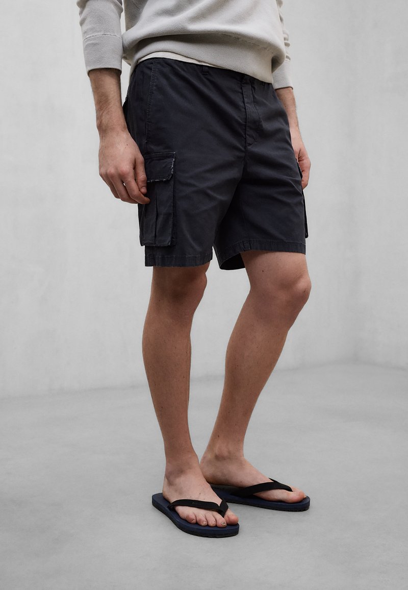Personne portant un short cargo noir et des tongs noires, debout sur un sol gris devant un mur uni gris clair.