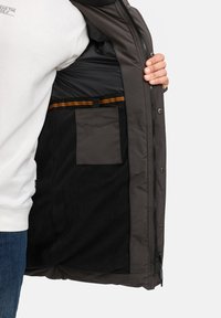 Giacca grigia con un morbido interno in pile nero. Presenta una tasca con zip e una striscia decorativa orizzontale. Chiusure con zip e bottoni automatici.