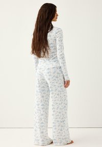 Γυναίκα με μακριά καστανά μαλλιά που φοράει λευκό ριμπ μπλουζάκι με λουλούδια και αντίστοιχο παντελόνι με φαρδιά μπατζάκια, όρθια ξυπόλυτη σε απλό φόντο.