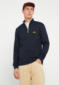 Marine blauwe sweatvest met ritssluiting en hoge kraag, met een geel berglogo op de borst, gedragen met lichtbeige broek.