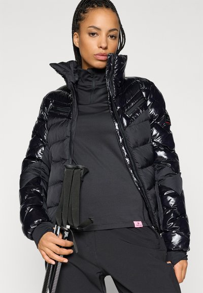 Casaco puffer preto com um acabamento brilhante, design acolchoado vertical, gola alta e fecho com zíper. A modelo segura varas de trekking pela cintura.