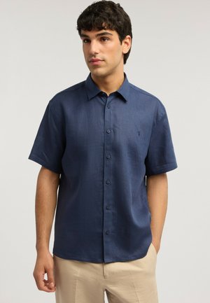 Junger Mann mit dunklem Haar, der ein marineblaues kurzärmeliges Hemd mit Knöpfen und beigen Hosen trägt und vor einem schlichten hellen Hintergrund steht.