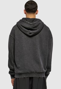 Melna kapuča krekls ar brīvu piegriezumu, izgatavots no teksturētas auduma, ar kangarū parādīšanās kabatu un ribotām aprocēm, skatīts no aizmugures.