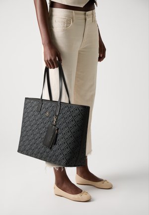 Black patterned tote bag with long handles and a small zip pouch attached. Smooth material with a rectangular shape. Worn with beige pants and flats.