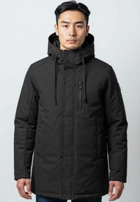 Homme portant un manteau d'hiver noir à capuche avec zip et fermetures à pression, se tenant contre un fond clair uniforme.