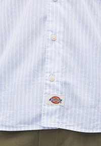 Λευκό πουκάμισο με λεπτές μπλε κάθετες ρίγες, δείχνει τρία κουμπιά και μια ετικέτα της μάρκας Dickies κοντά στο κάτω μέρος του ρεβέρ.