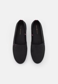 Ein Paar schwarze Slip-On-Canvas-Schuhe mit weißen Sohlen und dezenten rot, weiß und blauen Tommy Hilfiger-Logos an den Seiten und Innensohlen.