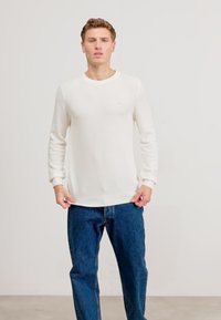 Witte katoenen trui met ronde hals en geribde manchetten en zoom, gecombineerd met blauwe spijkerbroek; model staat voor een neutrale achtergrond.