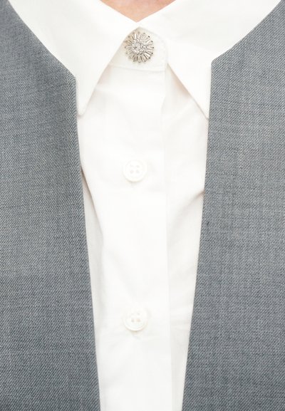 Λευκό πουκάμισο με ασημί φλοράλ καρφίτσα στο γιακά, φορεμένο κάτω από γκρι σακάκι με υφή.