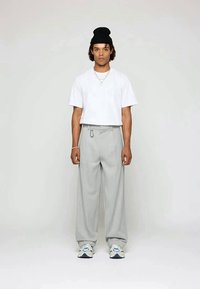 Young man wearing a white t-shirt, gray wide-leg pants, white sneakers, a black beanie, and silver necklaces standing against a plain background.