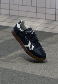 Marinblå sneaker i syntetmaterial med vita accenter, snörningsdesign och brun gummisula. Har en slät yta och varumärketing.