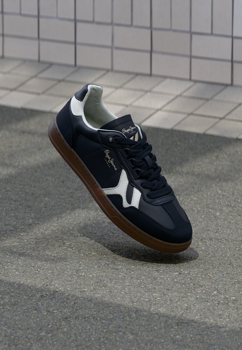 Marinblå sneaker i syntetmaterial med vita accenter, snörningsdesign och brun gummisula. Har en slät yta och varumärketing.