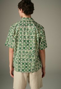 Chemise à manches courtes avec un motif géométrique vert sur fond beige. Fabriquée en tissu léger avec une coupe décontractée.