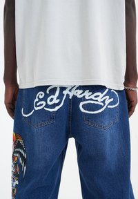 Jeansy dżinsowe w niebieskim odcieniu, z białym napisem "Ed Hardy" przeszytym na plecach. Misterny wzór czaszki z skrzydłami w żywych kolorach.