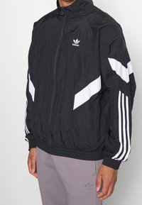 Svart vindjacka med vita diagonala ränder och Adidas-logotypen på bröstet. Har en ståkrage och elastiska manschetter.