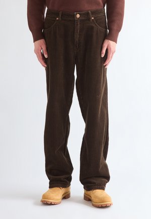 Man wearing dark brown corduroy pants, tan work boots, and a brown long-sleeve sweater, standing against a plain light background.