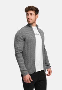 Grå strikket cardigan med ribbet tekstur, glidelås foran og ståkrage, brukt over en hvit skjorte. Har lange ermer og en tettsittende design.