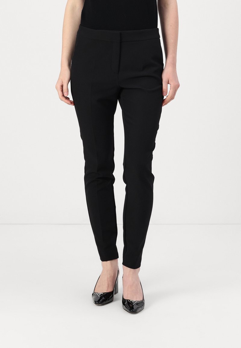 Femme debout portant un pantalon noir ajusté et des chaussures à talons hauts en cuir verni noir, sur un fond blanc.
