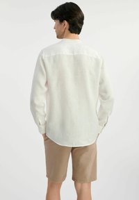 Lekka, lniana koszula z długim rękawem w kolorze off-white, z okrągłym kołnierzykiem i rozcięciami na mankietach, w połączeniu z beżowymi szortami. Gładka tekstura.