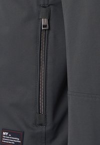 Primer plano de una cremallera negra vertical sobre tela gris oscura con un pequeño parche rectangular que presenta texto en blanco y naranja.