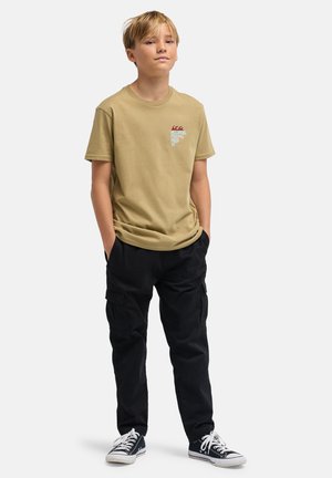 Khakifarbenes Baumwoll-T-Shirt mit einem Grafikdruck auf der Brust, kombiniert mit schwarzen Cargo-Hosen und schwarzen Turnschuhen mit weißen Schnürsenkeln.