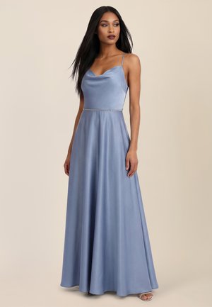 Femme portant une robe longue en satin bleu clair avec de fines bretelles et un détail de taille orné de perles, debout devant un fond beige uni.