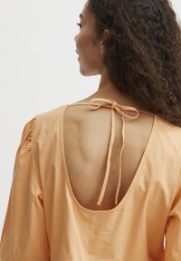 Perzikkleurige blouse met een diepe ruguitsnijding, afgewerkt met een bijpassende strik. Heeft pofmouwen en een zachte, gladde textuur.
