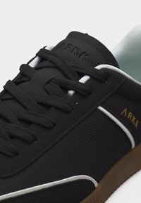 Μαύρο αθλητικό παπούτσι ARKK με μαύρα κορδόνια, λευκή εσωτερική επένδυση, λευκή επένδυση και καφέ σόλα, εμφανιζόμενο σε κοντινό πλάνο.