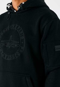 Primer plano de una persona que lleva una sudadera con capucha negra con el logo circular "Project X Paris" en el pecho y un pequeño parche en la manga.