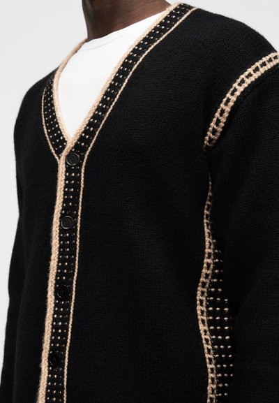 Μαύρη πλεκτή ζακέτα με μπεζ και μαύρη καρό διακόσμηση κατά μήκος του V-λαιμού, της γραμμής κουμπιών, του ώμου και της πλευράς, φορεμένη πάνω από λευκό πουκάμισο.