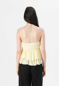 Top halter galben deschis cu un corset smockat și volane suprapuse la margine, având un șnur la gât și o textură moale și fluidă.