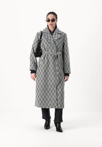 Cappotto lungo in pied-de-poule bianco e nero, con cintura in vita, colletto a risvolto e spalle cadenti. Indossato con jeans neri e stivaletti.
