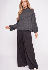 Szary sweter o fakturze z wysokim kołnierzem i szerokimi rękawami, zestawiony z czarnymi plisowanymi spodniami o szerokich nogawkach. Całość dopełniają białe buty.
