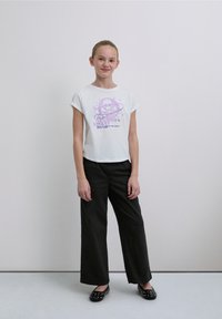 Tricou alb scurt cu un design grafic violet cu un craniu și text, asortat cu pantaloni negri evazați și balerini negri cu ținte.