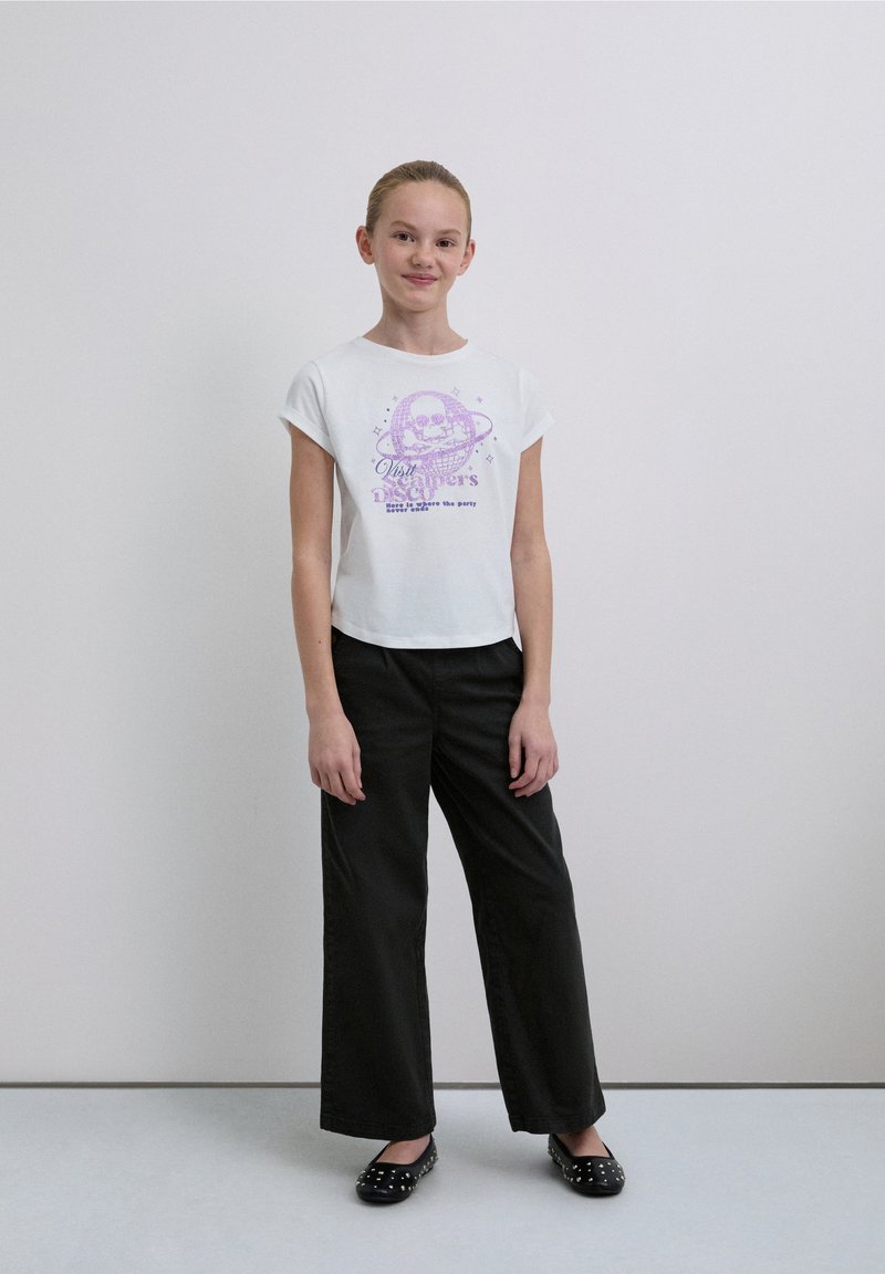 Tricou alb scurt cu un design grafic violet cu un craniu și text, asortat cu pantaloni negri evazați și balerini negri cu ținte.