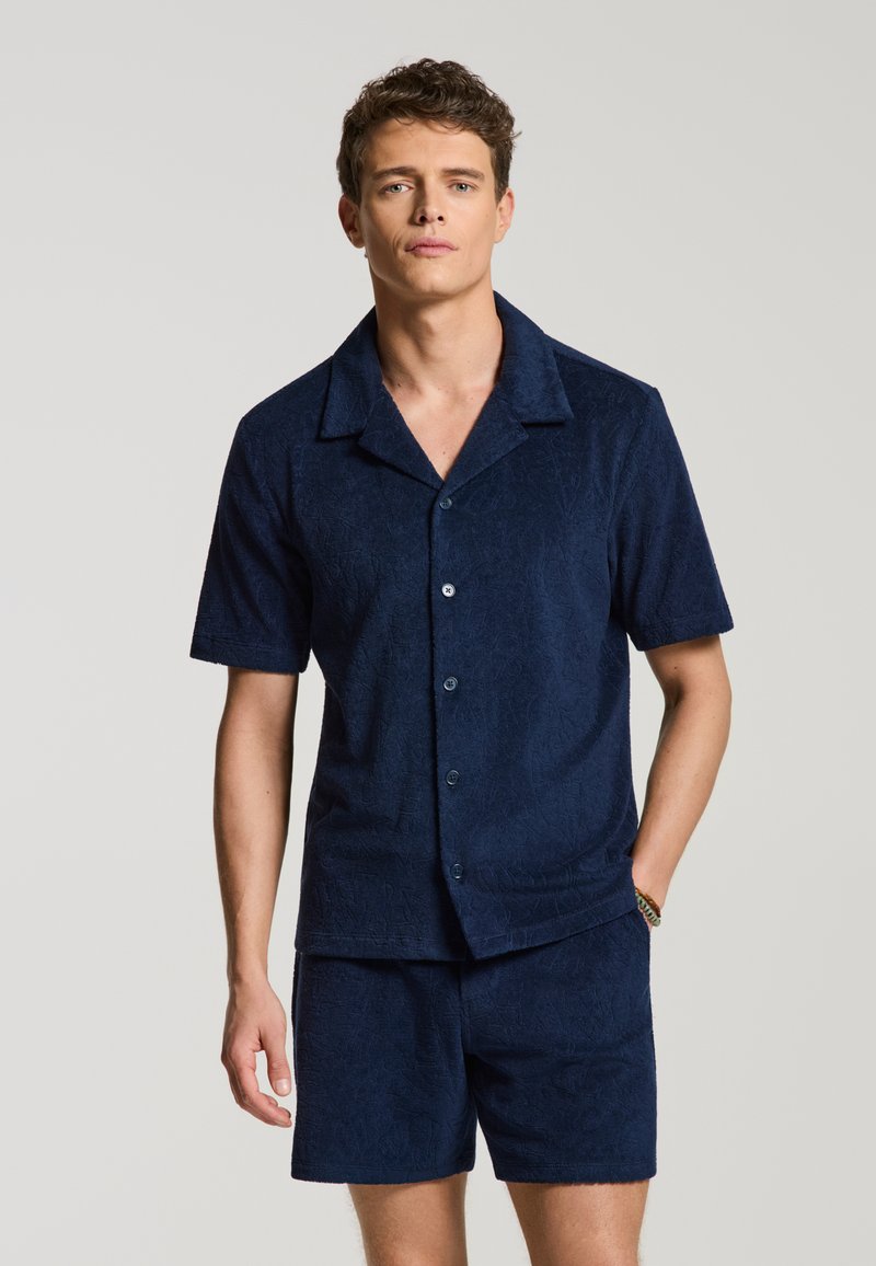 Junger Mann trägt ein marineblaues kurzärmeliges Hemd mit Knöpfen und dazu passende Shorts mit dezentem strukturiertem Muster, steht vor schlichtem Hintergrund.