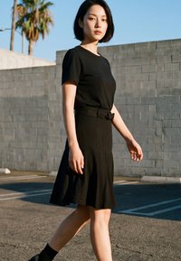 Young woman with short black hair wearing a black dress walks in an outdoor parking lot beside a gray concrete block wall and palm trees.