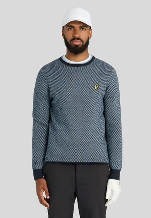 Mand iført en tekstureret blå sweater, sorte bukser, hvid golfhandske på højre hånd og hvid perforeret kasket, stående mod en ensfarvet baggrund.