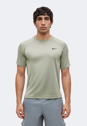 Jeune homme portant un t-shirt Nike à manches courtes vert clair et un short Nike gris, debout devant un fond uni.