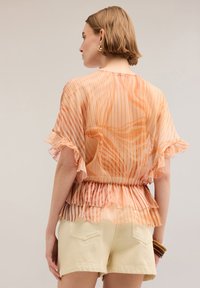 Blusa leggera e trasparente con strisce verticali arancioni e pesca, maniche a volant e vita arricciata, abbinata a pantaloni corti color crema.